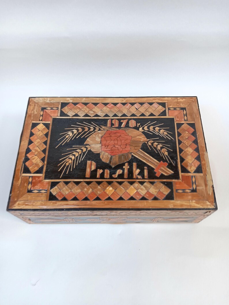 May include: A wooden box with a black background and a red flower design. The box is decorated with a geometric pattern of wood pieces. The text "1970r. Prustki" is visible on the box.