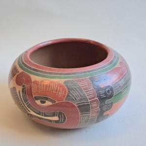 May include: A round, hand-painted ceramic bowl with a reddish-brown interior and a decorative exterior. The exterior features a design with red, black, green, and cream colours. The bowl has a wide opening and a rounded shape.