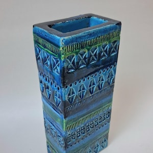 May include: A rectangular ceramic vase featuring a deep blue glaze with accents of lighter blue and green. The surface is decorated with horizontal bands of embossed geometric patterns, including diamonds, vertical lines, and small circular motifs.