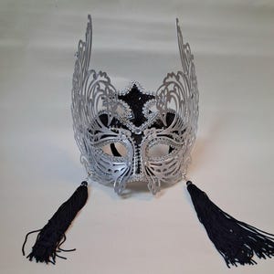 May include: Silver masquerade mask with an ornate filigree design, black detailing, and black tassels. The mask has a decorative crown and is embellished with rhinestones. The mask is suitable for a formal event or costume party.