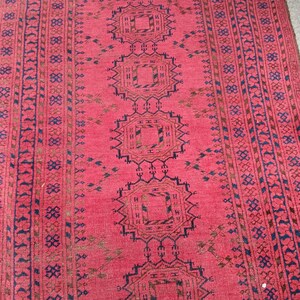 May include: A red oriental rug with a repeating geometric pattern in navy blue and brown. The rug features a central design of connected medallions and borders with intricate details. The rug has a fringe on one end.