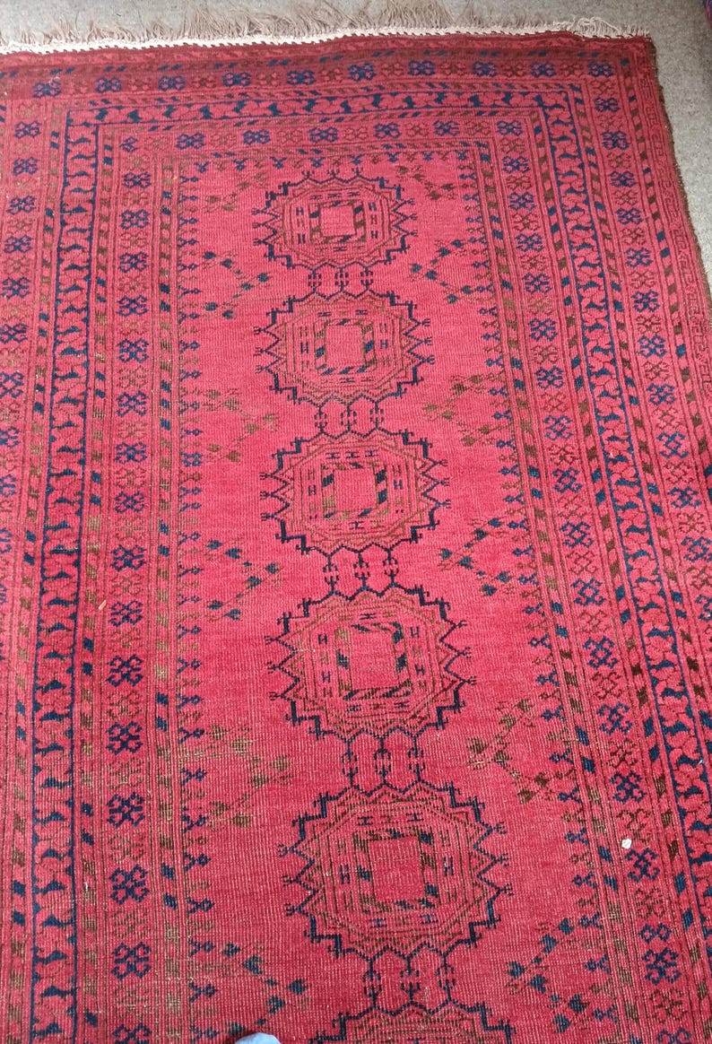 May include: A red oriental rug with a repeating geometric pattern in navy blue and brown. The rug features a central design of connected medallions and borders with intricate details. The rug has a fringe on one end.