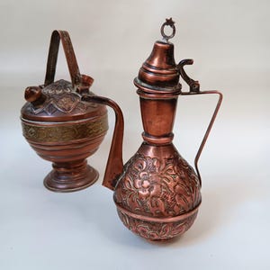 May include: Two antique copper teapots with intricate floral designs. The teapot on the left has a handle and a lid, while the teapot on the right has a spout and a lid.