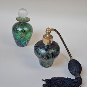 May include: Two antique glass perfume bottles. One is green with a frosted glass stopper. The other is dark blue with a gold-colored atomizer and a black tassel. Both bottles have a unique iridescent finish.