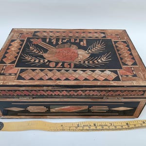 May include: A wooden box with a black background and intricate inlaid designs in shades of brown and red. The top of the box features a stylized image of a bird with wheat stalks and the text "1920" and "PRISTI".