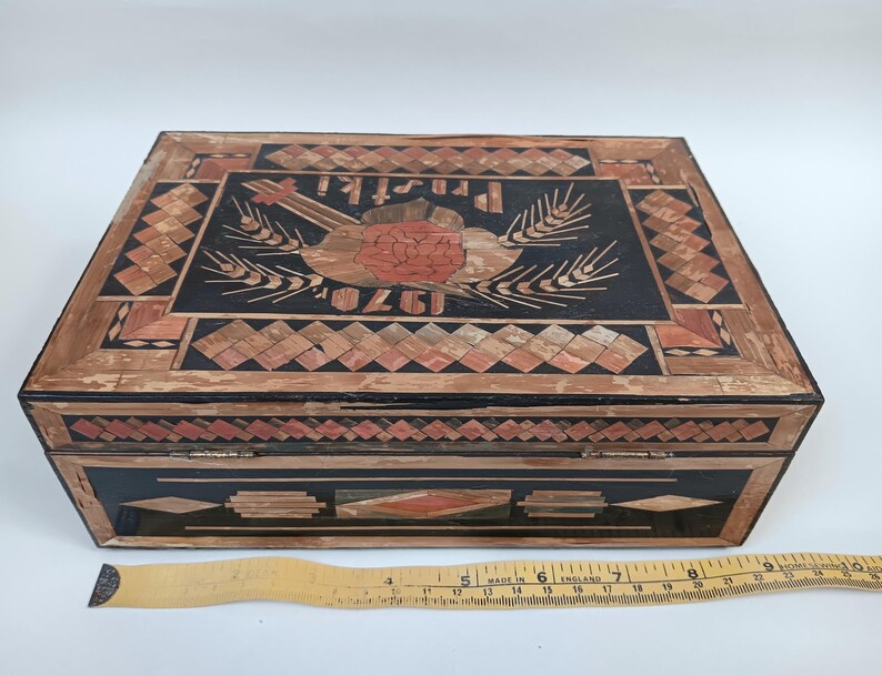 May include: A wooden box with a black background and intricate inlaid designs in shades of brown and red. The top of the box features a stylized image of a bird with wheat stalks and the text "1920" and "PRISTI".