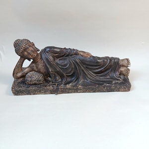 May include: A dark brown reclining Buddha statue. The figure is lying on its side, with one arm supporting the head. The statue is on a rectangular base. The Buddha is wearing a draped robe. The statue is approximately 30 cm long.