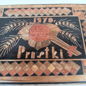 May include: A black wooden plaque with a straw art design. The design features a red flower with wheat stalks on either side. The plaque also includes the text "1970 r." and "Prustki".