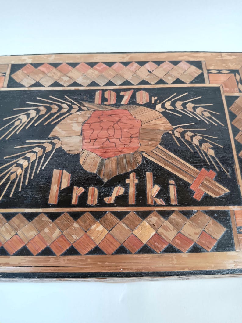 May include: A black wooden plaque with a straw art design. The design features a red flower with wheat stalks on either side. The plaque also includes the text "1970 r." and "Prustki".