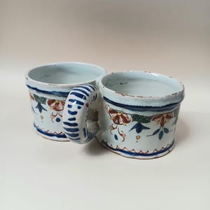 Könnte beinhalten: Ein Paar antike Keramikbecher mit einem gemeinsamen Henkel. Die Becher sind mit blauen, grünen und roten Blumenmustern auf weißem Hintergrund verziert.