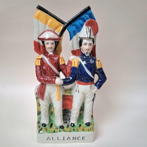 Puede incluir: Una figura de cerámica que representa a dos soldados con uniformes históricos, sosteniendo banderas. Un soldado viste un abrigo rojo, el otro un abrigo azul. Las banderas son amarillas, negras y azules. La base de la figura dice "ALLIANCE".