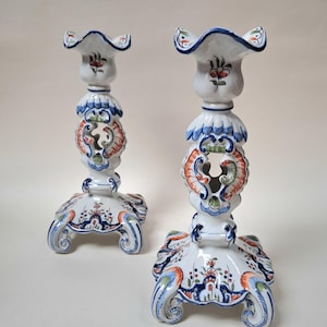 May include: Pair of white ceramic candlesticks with intricate floral and scroll designs in blue, orange, and green. Each candlestick features a scalloped top and a square base with curved feet. The overall height is approximately 8 inches.