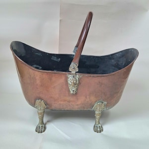 May include: A copper coal scuttle shaped like a miniature bathtub with a dark brown wooden handle. The vessel features brass lion head mounts where the handle attaches and stands on four ornate brass claw feet with a dark interior finish.