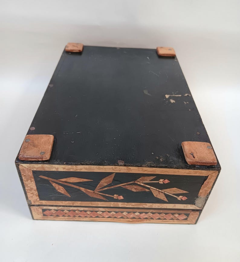 May include: A black wooden box with a floral design inlaid in wood. The box has a hinged lid and four small wooden feet.