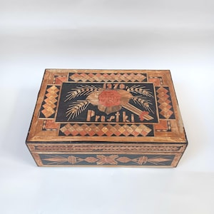 May include: A wooden box with a black background and intricate straw inlay. The top of the box features a red flower with wheat stalks and the text "1970r. Prustki".
