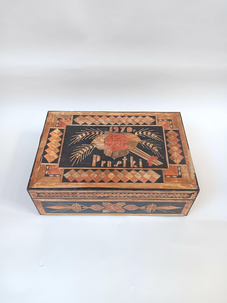 May include: A wooden box with a black background and intricate straw inlay. The top of the box features a red flower with wheat stalks and the text "1970r. Prustki".