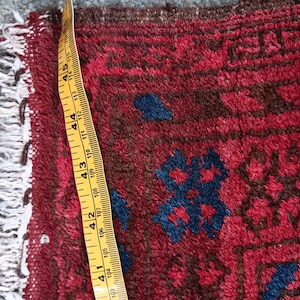 May include: Close-up of a section of a red and blue patterned rug. The rug has a geometric design with dark blue and brown accents. A yellow measuring tape shows a width of approximately 4 inches.