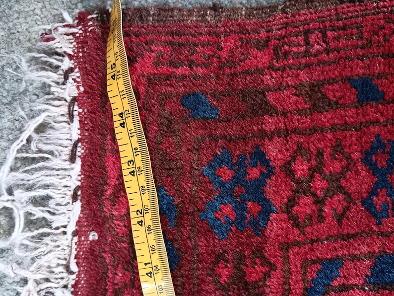 May include: Close-up of a section of a red and blue patterned rug. The rug has a geometric design with dark blue and brown accents. A yellow measuring tape shows a width of approximately 4 inches.