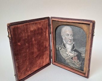 May include: A hinged, rectangular case with a dark brown exterior and a deep red velvet interior lining. The right side contains a portrait of a person in a military uniform with a sash and medals, framed by a gold-colored metal arch.