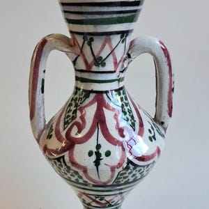May include: A decorative ceramic vase with two handles, featuring a white base with red, green, and blue painted patterns. The vase has a ruffled rim and a pedestal base, showcasing a traditional design.
