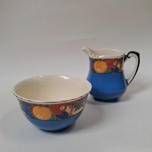 May include: A ceramic creamer and sugar bowl set featuring a vibrant blue base and a decorative band of stylised orange, red, and blue fruit motifs. Both pieces have a cream-coloured interior and a delicate gold-toned rim along the top edge.