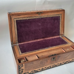 Könnte beinhalten: Eine rechteckige Schreibschatulle aus Holz mit Klappdeckel, die an den Rändern mit dunklen Holzeinlagen verziert ist. Die Innenseite des Deckels ist mit violettem Samt ausgekleidet, und der Boden enthält offene Fächer sowie ein Schlüsselloch.