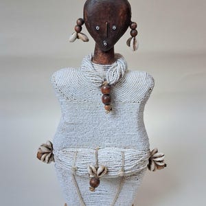 May include: A handcrafted sculpture of a figure with a dark wooden head and a body covered in white beads. The head is adorned with seashells and the figure has a necklace of wooden beads. The sculpture is mounted on a wooden base.