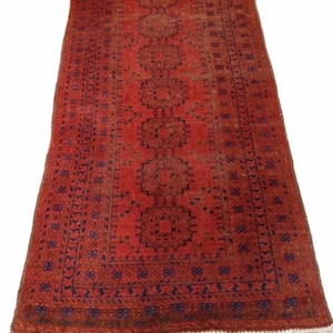 May include: A rectangular, red oriental rug with a central geometric pattern and a dark blue border. The rug has a distressed look, with visible wear and tear. White fringe edges the rug.