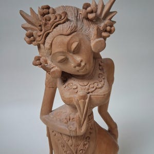 Könnte beinhalten: Eine hellbraune, handgeschnitzte Holzskulptur einer Figur mit geschlossenen Augen, die einen verzierten Kopfschmuck mit floralen Details, große kreisförmige Ohrringe und ein Oberteil mit komplizierten Mustern trägt, in einer anmutigen Pose.