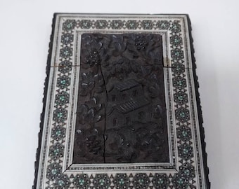 May include: A rectangular, dark wooden box with intricate carvings. The center features a detailed relief of a building and foliage. The border is inlaid with white and green decorative elements, creating a contrasting frame.