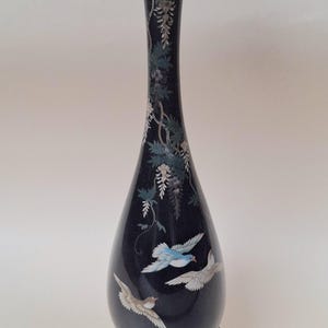 Pode incluir: Um vaso preto alto e esguio com pescoço cônico e base bulbosa. A superfície é decorada com ilustrações pintadas de três pássaros em voo e trepadeiras florais pendentes em tons de branco, azul e verde. Repousa sobre uma base de madeira escura.
