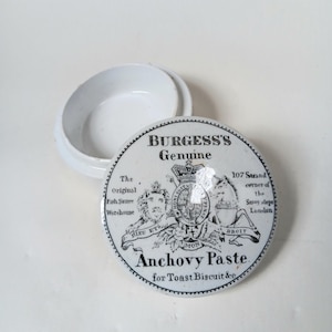 Puede incluir: Un recipiente de cerámica blanco y redondo con tapa. La tapa presenta el texto "BURGESS'S Genuine Anchovy Paste" con elementos decorativos y texto. El recipiente está abierto, revelando el interior.