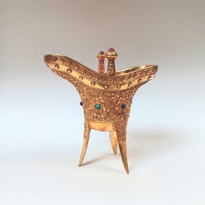 Puede incluir: Una copa de metal dorado con diseños intrincados y piedras preciosas azules y rojas. La copa tiene tres patas y un borde ancho y curvo.