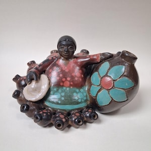 Puede incluir: Escultura de cerámica artesanal que representa una figura con un atuendo rojo y turquesa, sosteniendo un plato blanco y apoyada en un recipiente marrón con un diseño floral turquesa. La escultura está rodeada de pequeñas formas bulbosas de color marrón oscuro.