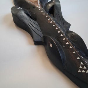 May include: A dark-stained wooden sculpture with white inlaid details. The artwork depicts a stylized bird or animal form with a light-coloured, textured section. The design incorporates geometric patterns and a flowing, curved shape.
