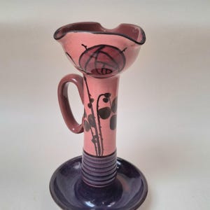 Könnte beinhalten: Ein Kerzenhalter aus Keramik mit einem rosafarbenen Oberteil, das mit einer handgemalten schwarzen Rose und einem Stiel verziert ist, einem dunkelvioletten Sockel mit horizontalen Rillen und einem geschwungenen Griff. Der obere Rand ist gewellt.