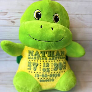 以下が含まれることがあります： 黄色の腹をした緑色のぬいぐるみのかめ。かめは刺繍された目と笑顔の口をしています。腹には「NATHAN MARTINS AZEVEDO」と「7 LB 12 OZ 20」、「12-03-2030」、「00:06 PM」の刺繍が施されています。