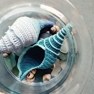Può includere: Due conchiglie all'uncinetto in una ciotola di vetro. La conchiglia più grande è blu chiaro e bianca, mentre la più piccola è blu scuro. Entrambe le conchiglie sono realizzate con un design testurizzato a spirale.