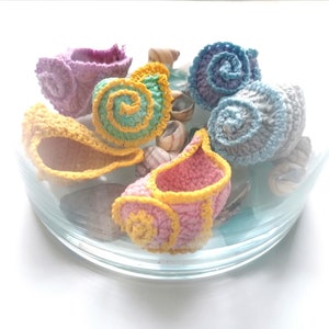 Peut inclure: Une collection de coquillages en crochet de différentes couleurs, dont le rose, le jaune, le bleu et le vert. Les coquillages sont disposés dans un bol en verre.