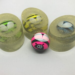 May include: Three clear, glittery soap bars with a bee design inside. A pink bee bouncy ball sits in front of the soap bars.
