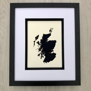 Puede incluir: Obra de arte enmarcada que presenta una silueta negra de Escocia sobre un fondo crema. La obra está enmarcada en un passe-partout blanco y un marco de madera negro. Una representación minimalista del paisaje escocés.