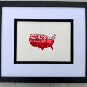 May include: Framed artwork featuring a red, textured depiction of the United States, crafted from a Coca-Cola can. The words "ORIGINAL TASTE" are visible. The artwork is set against a white mat and framed in a black frame.