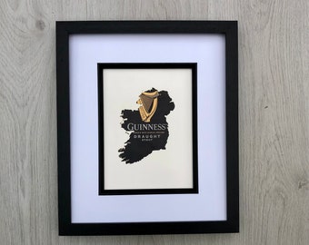 May include: A framed print featuring the Guinness logo and the outline of Ireland. The black frame surrounds a white matting, which highlights the black and gold design. The Guinness logo and text are visible.