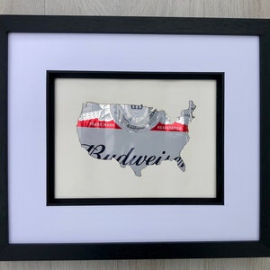 May include: Framed artwork featuring a map of the United States crafted from a Budweiser beer can. The artwork is set against a white mat and framed in a black wooden frame. The Budweiser logo is visible.