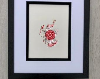 May include: Framed artwork featuring a map of Scotland in red and silver foil. The map is centered on a light background, framed with a white mat and a black wooden frame. The artwork includes text within a circular emblem.