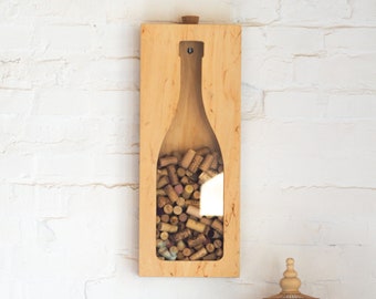 May include: A wooden wall mounted wine cork holder shaped like a wine bottle, filled with wine corks.  A wicker basket, a white ceramic vase, a ceramic pitcher, a birdcage, and a ceramic jar are on a wooden table in front of the cork holder.