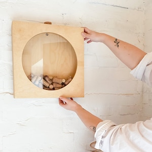 Könnte beinhalten: Ein wandmontierter Korkhalter aus Holz mit einer runden Öffnung und einem transparenten Glaseinsatz. Der Halter ist mit Weinkorken gefüllt.