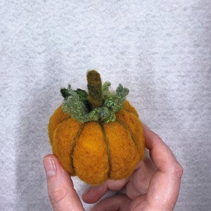 以下が含まれることがあります： 緑色の茎と葉が付いた、手作りの小さなフェルト製かぼちゃ。かぼちゃはオレンジ色で、表面がざらざらしています。