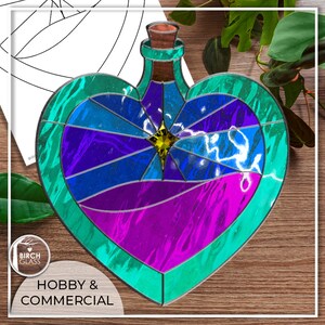 May include: A stained glass heart-shaped bottle with a blue, purple and green design. The bottle has a gold star in the centre and a clear glass stopper. The bottle is on a wooden surface.