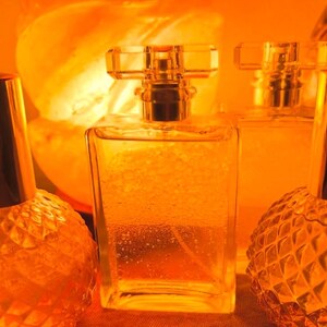 May include: Three glass perfume bottles with gold accents. The bottles are filled with a clear liquid and have a textured surface. The bottles are illuminated by a warm orange light.